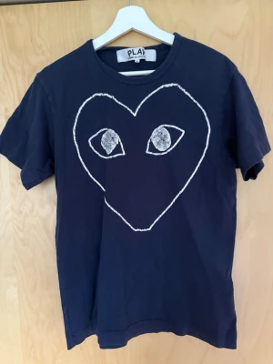 Mörkblå Comme des Garçons PLAY t-shirt - Säljer en mörkblå t-shirt från Comme des Garçons PLAY med ett stort vitt hjärta med ögon tryckt på bröstet. Klassisk rund hals och korta ärmar. Tillverkad i mjuk bomull som känns skön mot huden. Perfekt för dig som gillar streetwear och coola prints. Står L men sitter Medium