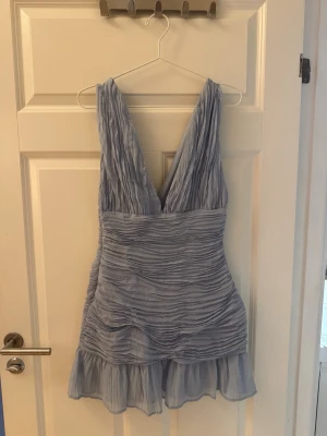 Ljusblå omlottklänning med volang - Supersöt ljusblå kortklänning med omlottdesign och volangkant. Klänningen har djup v-ringning och är ärmlös, med ett skirt, plisserat tyg som ger en luftig känsla. Perfekt för sommarens alla tillfällen!