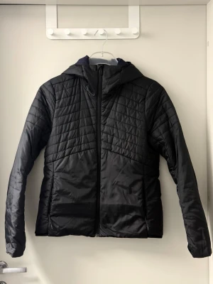Adidas svart vindjacka  - Snygg svart vindjacka med quiltade detaljer och huva. Jackan har dragkedja framtill och två sidofickor. Materialet är glansigt och lätt, perfekt för blåsiga dagar. Passar dig som gillar en stilren och sportig look.