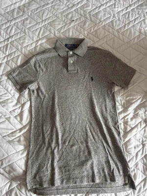 Grå pikétröja från Polo Ralph Lauren - Snygg grå pikétröja från Polo Ralph Lauren i slim fit-modell. Tröjan har klassisk krage, knappar framtill och den ikoniska broderade loggan på bröstet. Perfekt för en clean och stilren look.