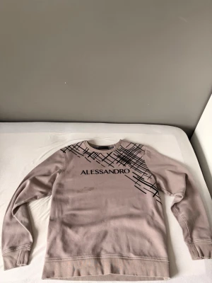 Beige sweatshirt från Alessandro Zavetti - Säljer en beige sweatshirt från Alessandro Zavetti med svart grafiskt tryck över bröstet och axeln. Tröjan har rund halsringning och långa ärmar. Materialet känns mjukt och bekvämt, perfekt för en avslappnad stil. Snygg detalj med loggan framtill.