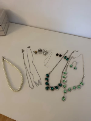 Mix av halsband, örhängen och ringar - Säljer ett paket med flera smycken: fem halsband, två med gröna stenar, ett pärlhalsband, två tunna silverfärgade kedjor samt örhängen i olika former och färger, bland annat gröna, svarta och silver. Perfekt för dig som vill variera din stil med olika accessoarer. Använda, säljer i pack