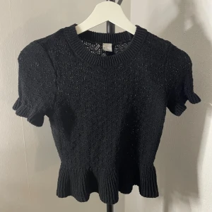 Svart stickad topp från H&M XS - Svart stickad topp från H&M i storlek XS. Toppen har korta ärmar, rund halsringning och en volangkant nedtill. Materialet är stickat och ger en mysig känsla, perfekt för att lyfta din outfit med en söt detalj.
