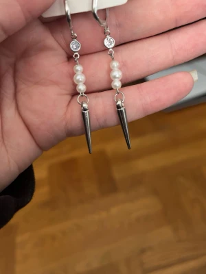 Hemma gjorda Silvriga örhängen med pärlor och spikes - Hemma gjordaSnygga örhängen i silverfärg med vita pärlor och spetsiga spikes längst ner. Örhängena har även en liten glittrande sten upptill. Perfekta för dig som gillar en edgy stil med en touch av elegans.