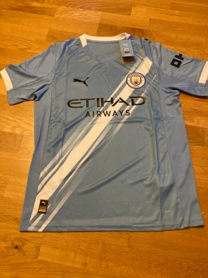 Manchester City matchtröja Puma - Snygg ljusblå Manchester City matchtröja från Puma med vita detaljer och klubbmärke på bröstet. Tröjan har svart Puma-logga, Etihad Airways-tryck och vita ränder diagonalt framtill. Tillverkad i lätt och ventilerande polyester, perfekt för fotboll eller träning.