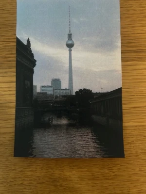 Fotoprint av Berlins TV-torn - Säljer en fotoprint med motiv av Berlins ikoniska TV-torn, Fernsehturm, med stadssilhuett och vatten i förgrunden. Bilden har en dämpad färgskala i grått och svart och är tryckt på matt papper. Perfekt för dig som gillar urbana motiv och vill ha något unikt på väggen.