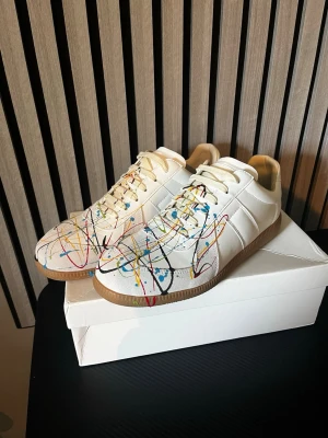 Maison margella - Unika vita sneakers med färgstänk i blått, rött, gult och svart. Skorna har klassisk låg silhuett, vita snören och beige gummisula. Tillverkade i slätt läder med en cool konstnärlig touch framtill. Perfekta för dig som vill sticka ut med din stil.