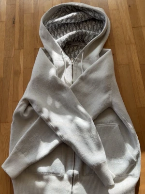 Vit hoodie från D I O R med monogram  - Vit hoodie från  D I O R med dragkedja och fickor framtill. Insidan av huvan har ett grått  D I O R-monogrammönster som ger en lyxig vibe. Tillverkad i mjukt bomullsmaterial och har broderad D I O R-logga på fickan. Perfekt för dig som gillar exklusiva streetwear-plagg.