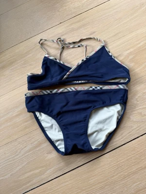 Burberry bikini 12y - Snygg mörkblå Burberry  bikini med omlottopp och smala axelband. Kanterna har ett diskret rutigt mönster i beige, svart och rosa som ger en unik touch. Perfekt för lata dagar på stranden eller poolen.