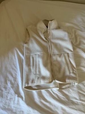 Beige fleeceväst med dragkedja - Mysig beige fleeceväst med hög krage och dragkedja framtill. Västen har två fickor på sidorna och är tillverkad i ett mjukt, fluffigt material som ger en avslappnad look. Perfekt att slänga över en hoodie eller tröja för extra värme.