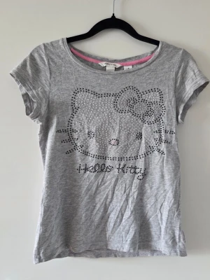 Grå Hello Kitty t-shirt med strass - Snygg grå t-shirt med korta ärmar och ett stort Hello Kitty-motiv framtill i glittriga strassdetaljer. Den är från barnavdelningen så ganska liten i storlek, skulle säga att storleken mostvarar xs ungefär