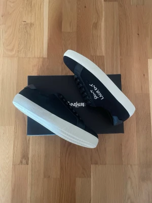 Svarta Saint Laurent sneakers - Svarta sneakers från Saint Laurent med vit sula och handskriven logga på sidan. Skorna har klassisk låg profil, svarta skosnören och är tillverkade i canvas med detaljer i läder. Perfekta för en clean och stilren look.