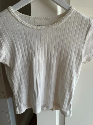 Vit ribbad topp med kort ärm - Säljer en vit ribbad topp med korta ärmar och rund hals. Toppen har en diskret struktur i tyget och är gjord i ett mjukt material som känns skönt mot huden. Perfekt att styla med jeans eller kjol för en clean look.