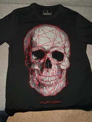 Svart Philipp Plein t-shirt med dödskalle - Snygg svart t-shirt från Philipp Plein med en stor dödskalle framtill i glittriga stenar och röda linjer. T-shirten har korta ärmar och rund hals. Märkesloggan är broderad i rött nedanför motivet. Perfekt för dig som gillar statement-plagg.