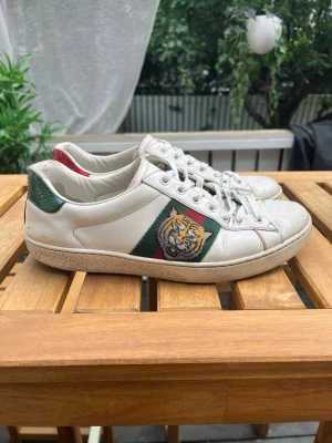 Zara - Gcci sneakers i vitt läder med klassiska gröna och röda ränder på sidan och en broderad tigerpatch. Baksidan har ormmönstrade detaljer i rött och grönt. Skorna har vita snören och en låg silhuett som ger en clean och lyxig vibe.