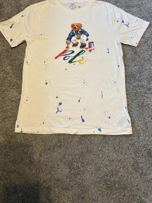 Vit Polo Ralph Lauren t-shirt med björn - Vit t-shirt från Polo Ralph Lauren med färgstänk och ikonisk björn på bröstet. Trycket visar björnen som målar ordet 'Polo' i flera färger. Klassisk passform och korta ärmar. Perfekt för dig som gillar streetwear och unika detaljer.