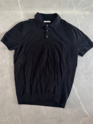 Svart polotopp från ZARA - Svart polotopp från ZARA med klassisk krage och tre knappar framtill. Toppen har korta ärmar och är tillverkad i ett mjukt material som känns skönt mot huden. Enkel och stilren design som passar till många olika outfits.