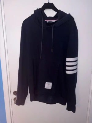 Thom browne liknande hoodie  - Snygg mörkblå hoodie från TBTHOM BULAN med huva och dragsnören. Har en stor magficka och tre vita ränder på ena ärmen. Detaljerad etikett på fickan och klassisk ribbad mudd vid ärmslut och nederkant. Perfekt för en avslappnad streetwear-look. Stl M men sitter bra på mig som är 175 och väger 60 kg 