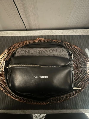 Svart handväska från Valentino - Snygg svart handväska från Valentino med silverfärgade detaljer och dragkedja. Väskan har ett stort fack och ett mindre ytterfack med logotypen tryckt på framsidan. Axelremmen är justerbar och väskan har en modern, stilren design med logomönster på ovansidan.