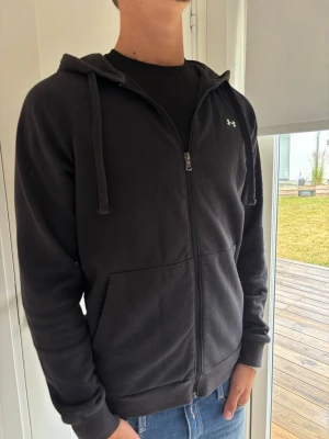 Svart zip hoodie från Under Armour - Säljer en svart hoodie från Under Armour med dragkedja framtill, två stora fickor och justerbar huva med snören. Klassisk logga på bröstet och ribbade muddar vid ärmar och nederkant. Perfekt för chill eller träning.