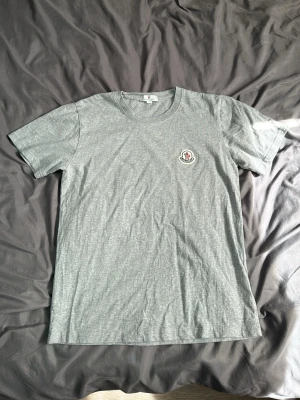 Grå Moncler t-shirt med logga - Snygg grå t-shirt från Moncler med klassisk rund hals och korta ärmar. På bröstet sitter Monclers ikoniska logga i vitt, rött och blått. Tillverkad i mjuk bomull som känns skön mot huden. Perfekt till jeans eller shorts för en clean look. Liten hål under armhålan men inget som syns. PASSAR M LIKA BRA SOM L.