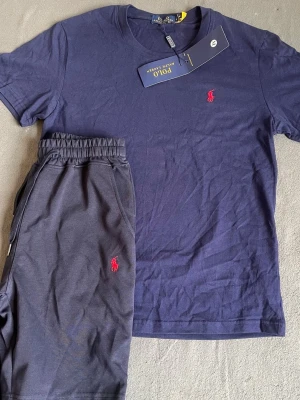 Mörkblått set från Polo Ralph Lauren - Snyggt mörkblått set från Polo Ralph Lauren med t-shirt och shorts. Båda plaggen har det klassiska röda Polo-logotrycket. T-shirten har rund hals och shortsen har resår i midjan för skön passform. Perfekt för en avslappnad och stilren look.