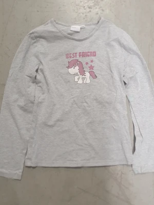 Tröja - Ljusgrå långärmad tröja med tryck av en söt enhörning och texten 'Best Friend' i rosa. Tröjan har rund halsringning och är gjord i mjukt bomullsmaterial. Perfekt för dig som gillar gulliga motiv och vill ha en bekväm vardagströja. Finns fläck.