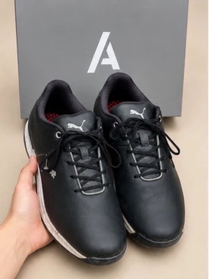 Puma Golfskor - Svarta golf skor från Puma med vita detaljer och logga på sidan. Skorna har lågt skaft, svarta skosnören och en vit sula med textur. Ovandelen är i slätt material och insidan har röd vaddering. Perfekta för en sportig och clean look.