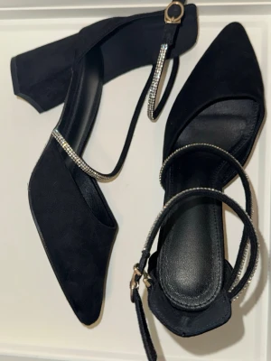 Svarta pumps med strassdetaljer - Snygga svarta pumps med spetsig tå och bred klack. Skorna har två tunna remmar med glittrande strass och spänne vid ankeln. Materialet ser ut som mockaimitation och innersulan är svart. 