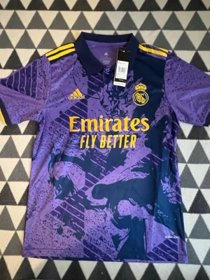 Lila Real Madrid matchtröja Adidas - Säljer en lila Real Madrid matchtröja från Adidas med gula detaljer och klubbmärke på bröstet. Tröjan har krage, korta ärmar och trycket 'Emirates Fly Better' på framsidan. Tillverkad i lätt och ventilerande material, perfekt för fotboll eller träning.