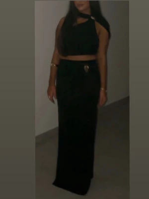 Svart tvådelat set med detaljer - Säljer ett svart tvådelat set med crop top och lång kjol. Toppen har asymmetrisk axel och dekorativa ringar vid axel och hals. Kjolen är figurnära och har en metallisk detalj i midjan. Perfekt för dig som vill ha en trendig och stilren look.