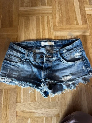 Blå jeansshorts med fransig kant - Säljer ett par blå jeansshorts från Zara med fransig kant och slitna detaljer. Shortsen har fem fickor, bälteshällor och knappgylf. Perfekta för varma dagar och ger en avslappnad vibe.