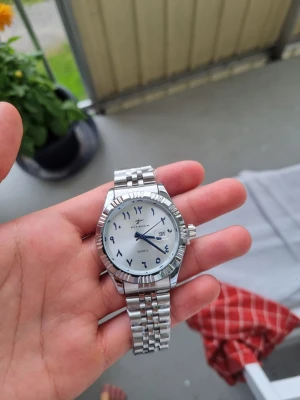 Silverfärgad klocka med arabiska siffror - Stilren klocka från ricecgo med silverfärgad länk i metall och vit urtavla. Urtavlan har arabiska siffror och datumvisning. Klassisk design med räfflad bezel och kvartsverk. Perfekt accessoar för dig som gillar en clean look. Helt ny aldrig använd ej kortad alla länkar i går och är fullt fungerande skulle kunna sänka priset vid snabbköp