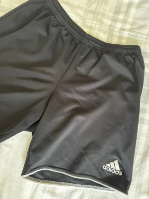 Svarta träningsshorts från Adidas - Säljer ett par svarta träningsshorts från Adidas med elastisk midja och diskret logga på benet. Shortsen är tillverkade i lätt och bekvämt syntetmaterial, perfekta för sport och aktivitet. Klassisk design med enkel passform och vit detalj vid benslut.