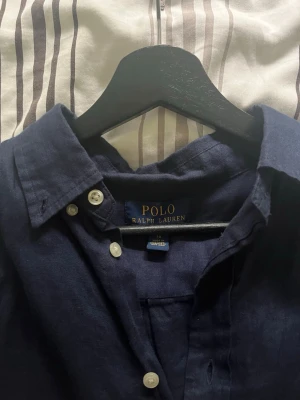 Mörkblå linneskjorta från Polo Ralph Lauren - Snygg mörkblå skjorta från Polo Ralph Lauren i linne. Klassisk modell med knappar framtill och lång ärm. Ikoniska röda loggan broderad på bröstet. Perfekt för en stilren och avslappnad look. Hör av er om det finns några frågor.