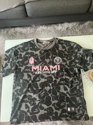Svart camo BAPE x Miami t-shirt - Säljer en svart och grå camo t-shirt från BAPE x Miami. Storlek L men passar även XL. Oanvänd och med taggarna kvar, kommer i original förpackning. Kan skicka fler bilder privat. 