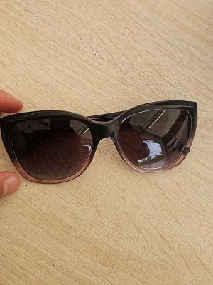 Svarta cat eye solglasögon Emporio Armani - Snygga cat eye solglasögon från Emporio Armani med svart båge som tonar ut i ljusrosa nedtill. Glasen är mörka och ger en cool look. Perfekta för dig som vill sticka ut med en trendig accessoar.
