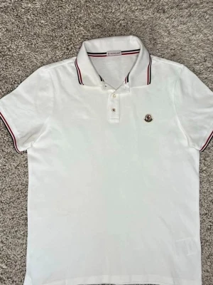 Vit Moncler pikétröja med röd detalj - Vit pikétröja från Moncler med klassisk krage och röda samt svarta ränder på krage och ärmslut. Tröjan har korta ärmar, knappar framtill och Moncler-logga på bröstet. Perfekt för en clean och sportig look.