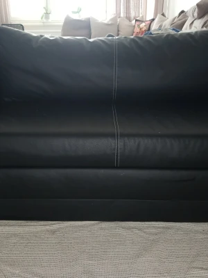 Svart bäddsoffa i konstläder - Säljer en svart bäddsoffa i konstläder med vita kontrastsömmar. Soffan kan enkelt fällas ut till en säng och har två armstöd samt en matchande kudde. Perfekt för dig som vill ha en smidig lösning för både sitt- och sovplats.         Läder soffa.      Soffan hämtas från Nybohovsbacken 87