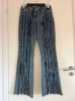 Blå jeans med fransade detaljer - Unika blå jeans med flera vertikala fransade och slitna detaljer längs hela benen. Modellen är rak med lätt utsvängda ben och hög midja. Jeansen har klassisk femficksdesign och stängs med knapp och dragkedja. Perfekta för dig som vill sticka ut med din stil.