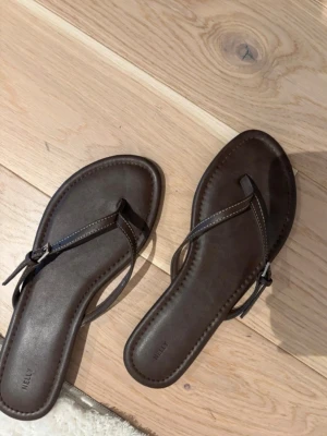 Bruna flip-flop sandaler från Nelly - Snygga bruna flip-flop sandaler från Nelly med tunna remmar och diskreta sömmar längs kanterna. Klassisk design med platt sula och tårem, perfekta för sommardagar. Tillverkade i ett material som ser ut som konstläder.