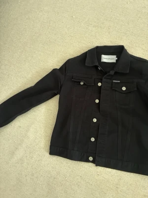 Svart jeansjacka från Calvin Klein - Snygg svart jeansjacka från Calvin Klein med klassisk krage, silverfärgade knappar och två bröstfickor med lock. Jackan har raka linjer och en stilren look som passar till det mesta. Perfekt för dig som gillar en enkel men trendig stil. Inga anmärkningar passar även M
