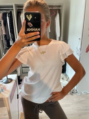 Vit ribbad topp med volangärm - Säljer en söt vit ribbad topp med korta volangärmar. Toppen har rund halsringning och är i ett mjukt, stretchigt material som sitter bekvämt. Perfekt att styla med jeans eller kjol för en fräsch look.