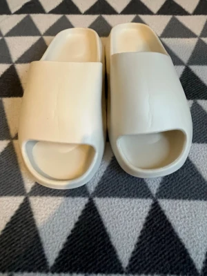 Beige chunky slidesandaler - Chunky slidesandaler i beige med öppen tå och bred rem över foten. Tillverkade i ett mjukt och flexibelt gummiliknande material som ger en clean och modern look. Perfekta för en avslappnad stil och enkel att matcha med olika outfits.