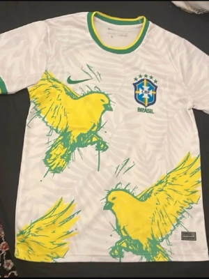 Vit Brasilien fotbollströja Nike - Säljer en vit Brasilien fotbollströja från Nike med gröna och gula detaljer. Tröjan har stora gula fåglar tryckta på framsidan, CBF-logga och Nike Swoosh på bröstet. Tillverkad i Dri-FIT material som andas, perfekt för träning eller match.