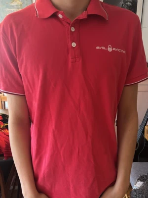 Röd Sail Racing piké t-shirt - Röd piké t-shirt från Sail Racing med vit logga på bröstet och vita detaljer på krage och ärmslut. Klassisk pikékrage med tre knappar framtill. Tillverkad i mjuk bomull och har en normal passform.