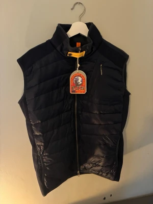 Mörkblå dunväst från Parajumpers - Snygg mörkblå dunväst från Parajumpers med hög krage och gul detalj vid halsen. Västen har dragkedja framtill, två sidofickor och en bröstficka med dragkedja. Tillverkad i ett glansigt material med quiltad design och logga på bröstet och nacken. - vid snabba köp kan priset sänkas🔥
