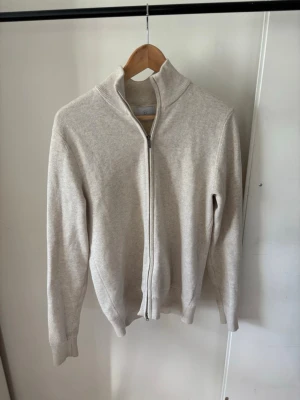 Beige ziptröja med hög krage - Snygg beige ziptröja med hög krage och långa ärmar. Tröjan har ribbade muddar vid ärmslut och nederkant samt dragkedja framtill. Perfekt för lager på lager och enkel att matcha med olika outfits.