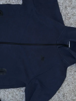 Mörkblå hoodie från Nike - Snygg mörkblå hoodie från Nike med dragkedja framtill och diskret Nike-logga på bröstet. Hoodien har huva och är tillverkad i mjukt bomullsmaterial som känns skönt mot huden. Perfekt för en avslappnad och sportig stil.