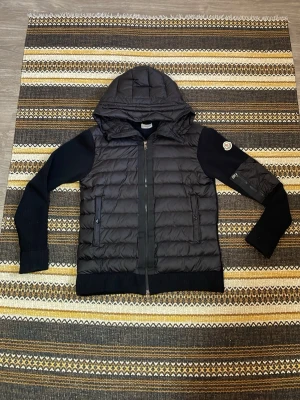 Moncler Cardigan  - Svart cardiganjacka från Moncler med quiltad front och huva. Ärmarna är ribbstickade och har en patch med Moncler-logga på vänster arm. Jackan har dragkedja framtill och två fickor med knappstängning. Materialmix av dun och stickat ger en modern look. Passar även storlek S för den passar mig ⭐️
